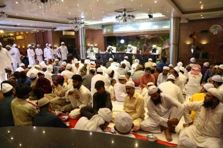 Wide view of a massive gathering ($\text{Ijtima}$) of Muslim attendees and students listening to the panel of scholars led by Shaykh Muhammad Mohsin Munawar Yousafi, seated on the floor.
