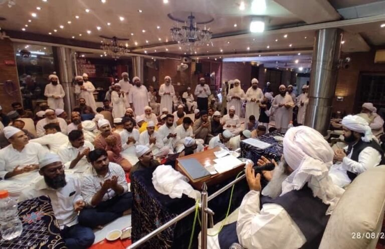 Shaykh Muhammad Mohsin Munawar Yousafi, the esteemed Islamic spiritual guide (Murshid), giving a discourse into a microphone, hands gesturing, wearing a white turban and dark vest.
