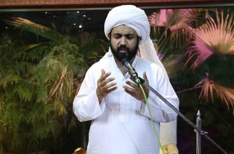 Shaykh Barrister Ahmad Mohsin Mohsini delivering a religious address (Sohbat) into a microphone, wearing a white turban, with hands raised in a gesture of spiritual guidance.
